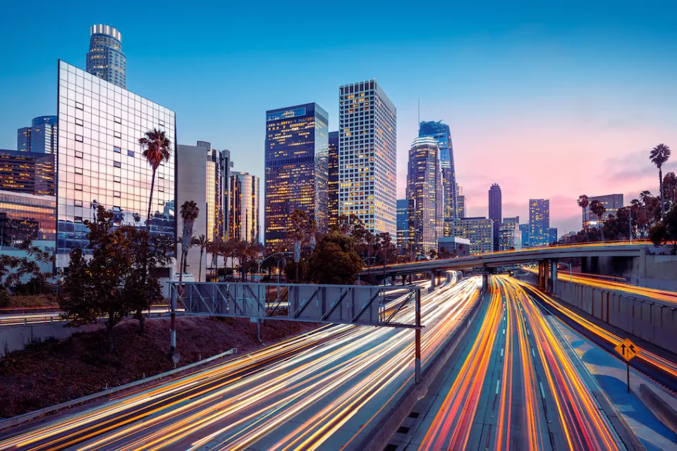 Blurred traffic speeding by on freeway with Los Angeles skyline in background.